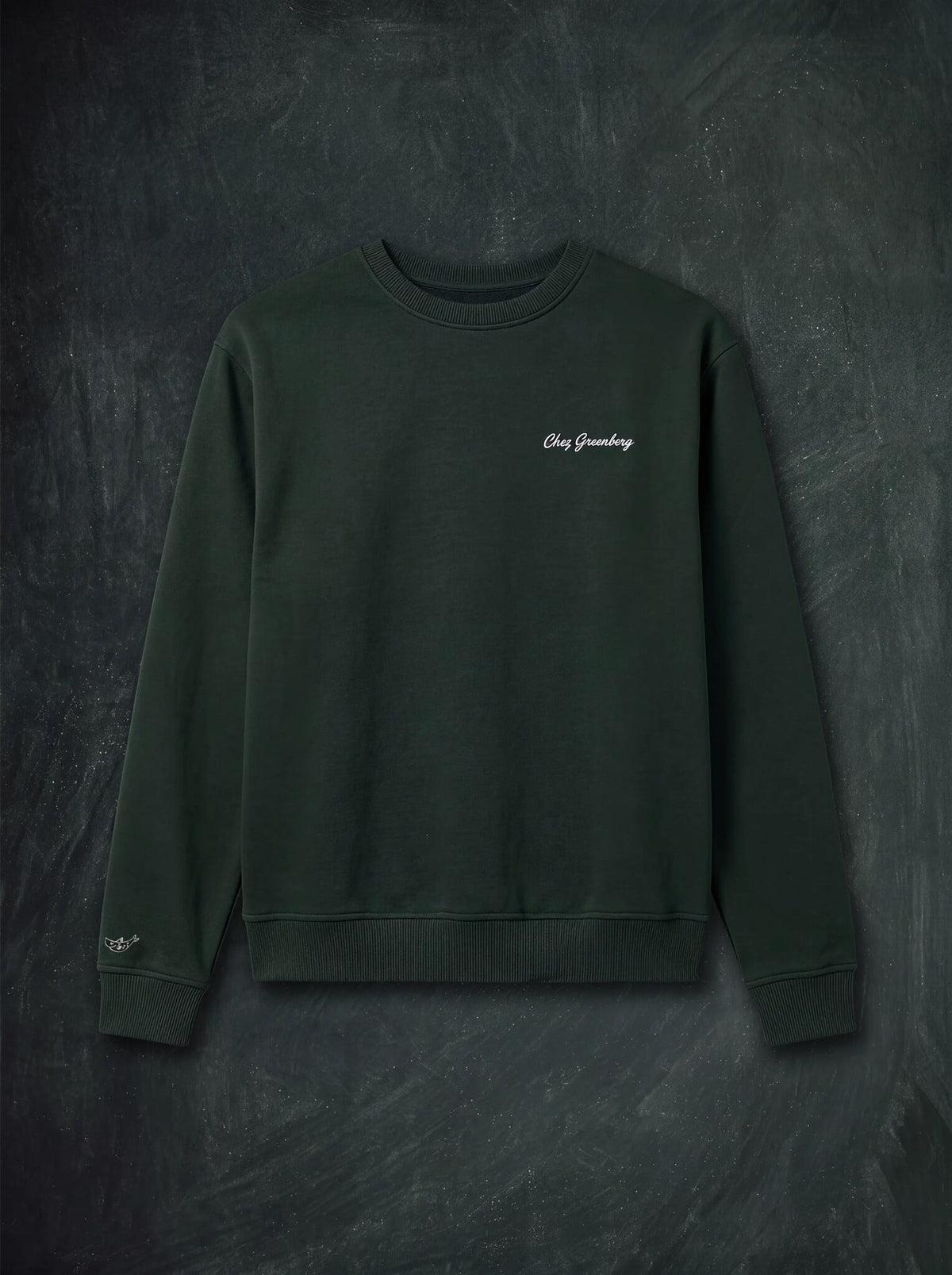 A dark forest green crewneck sweatshirt with 'Chez Greenberg' embroidered in cream script on the left chest, displayed flat against a dark textured background.