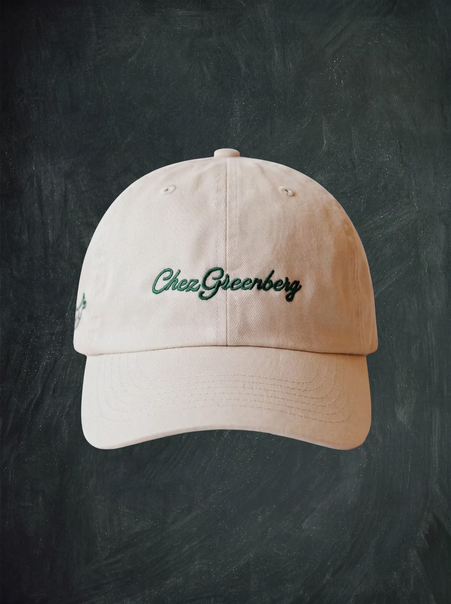 A light beige baseball cap with 'Chez Greenberg' embroidered in dark green script on the front, displayed against a dark textured chalkboard background.