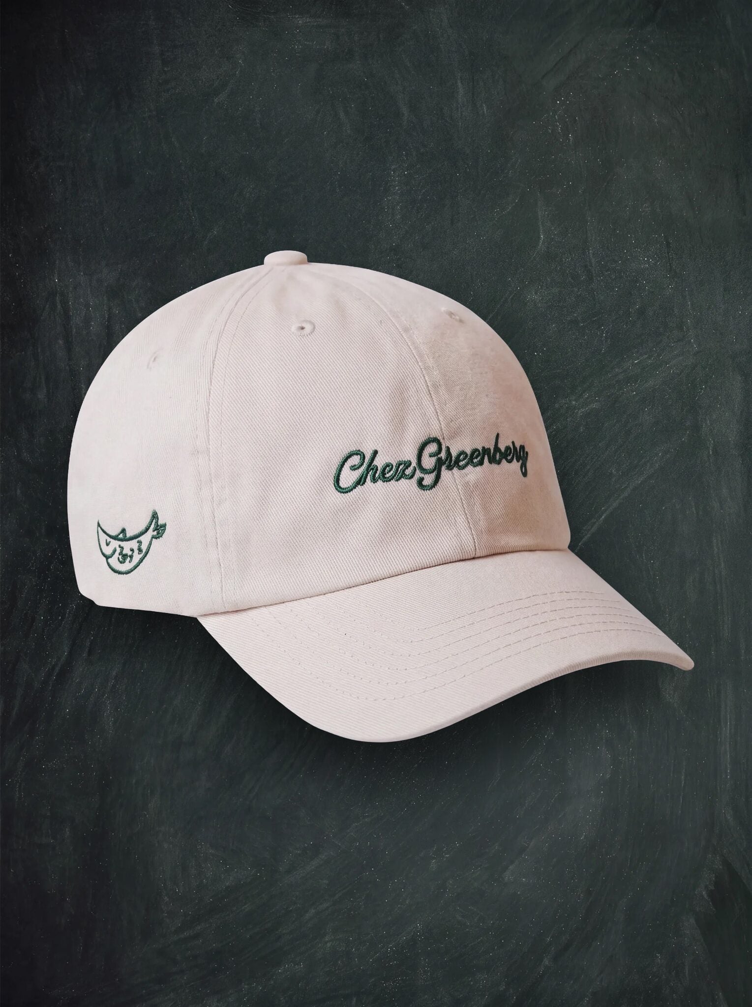 A light cream baseball cap with 'Chez Greenberg' embroidered in dark green on the front and a small matching green logo on the side, displayed against a dark chalkboard background.