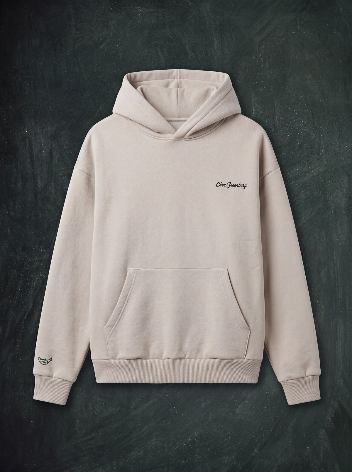 An icy-beige heavy-weight pullover hoodie with a small green script logo on the left chest and a small embroidered detail on the right sleeve, displayed flat against a dark textured background.
