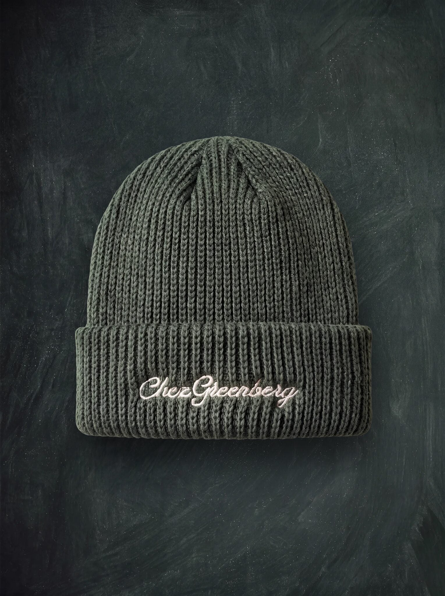 A green ribbed knit beanie with 'Chez Greenberg' embroidered in white script on the front cuff, displayed against a dark textured chalkboard background.