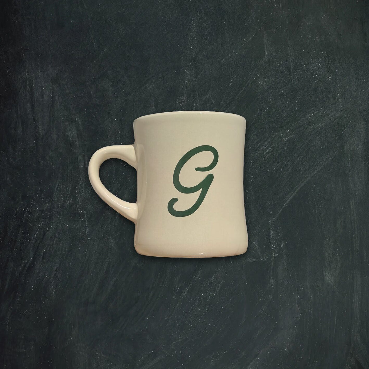 An off-white ceramic mug with a dark green cursive letter 'G' printed on the side, set against a dark textured chalkboard background.
