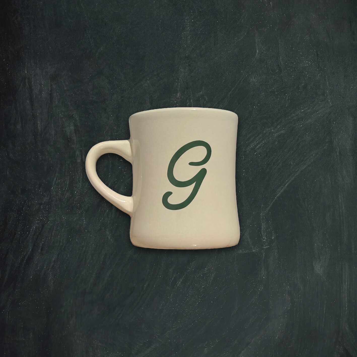 An off-white ceramic mug with a dark green cursive letter 'G' printed on the side, set against a dark textured chalkboard background.