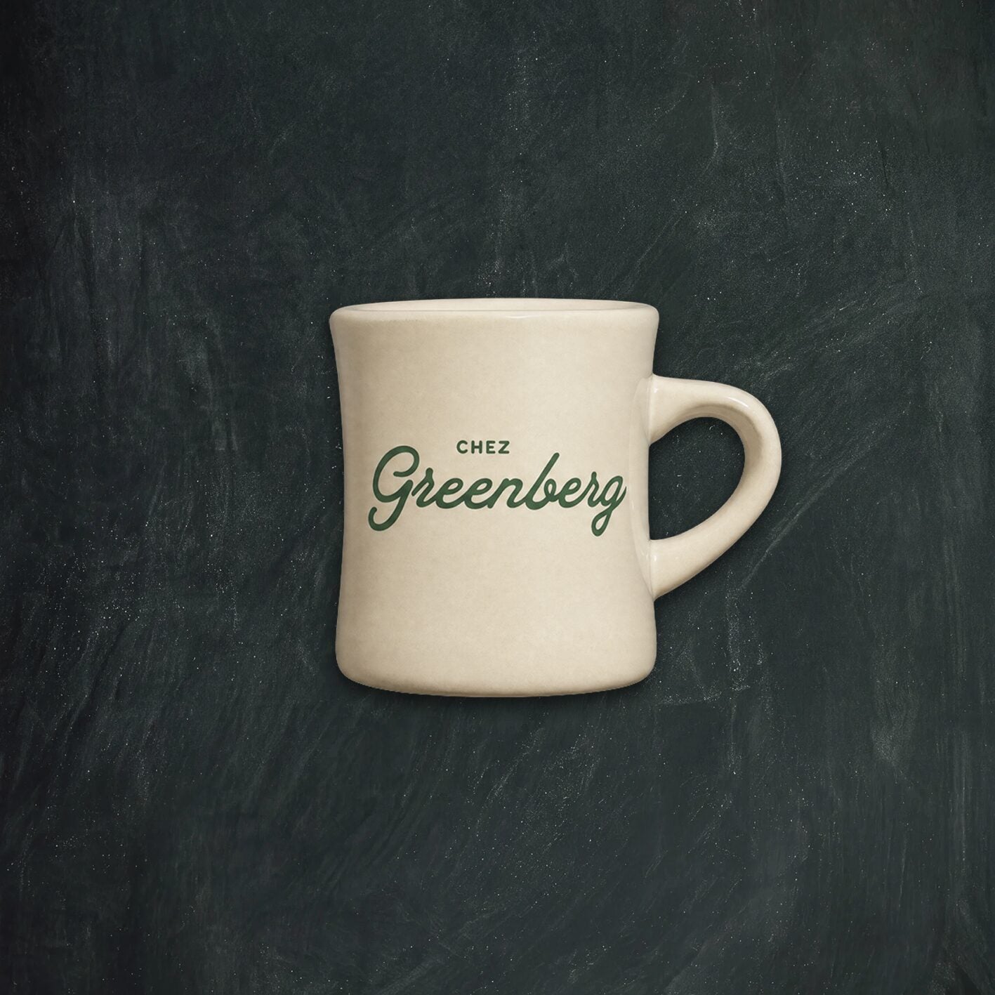 A cream-colored ceramic mug with 'CHEZ Greenberg' printed in dark green script on the front, set against a dark textured chalkboard background.