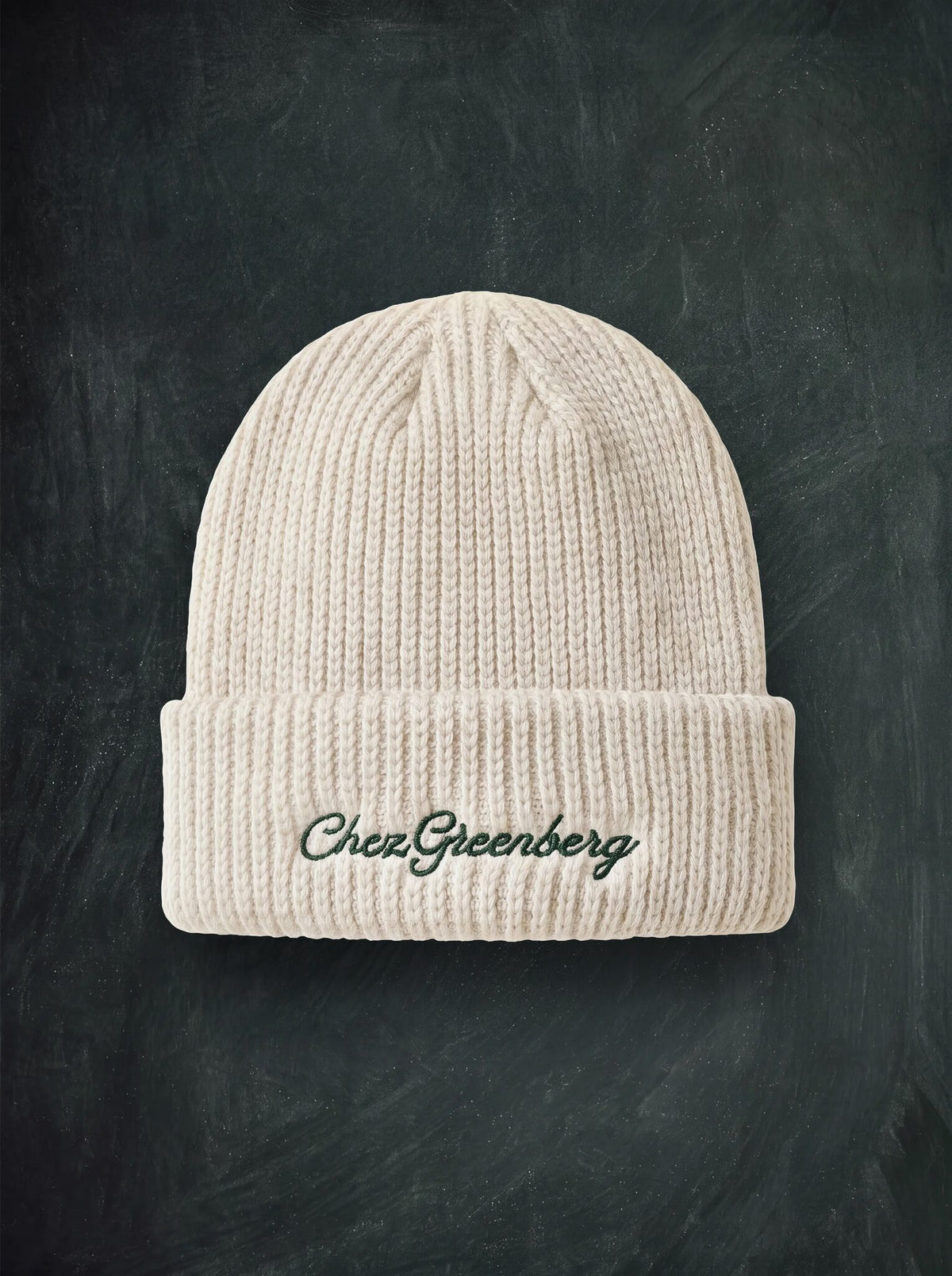 A cream-colored ribbed knit beanie hat with 'Chez Greenberg' embroidered in dark green script on the front cuff, displayed against a dark chalkboard background.