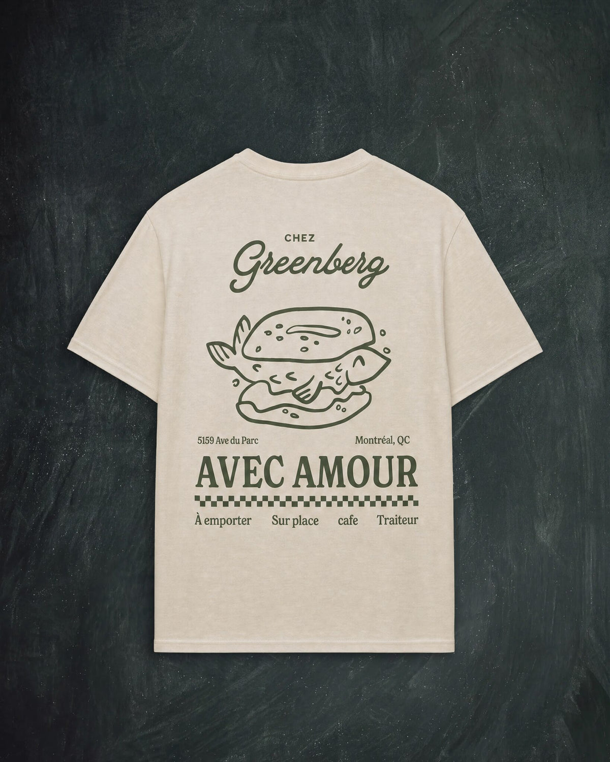 An off-white short-sleeve t-shirt with a dark green graphic print on the back, featuring a smoked salmon bagel illustration and the text 'CHEZ Greenberg' and 'AVEC AMOUR', displayed against a dark chalkboard background.